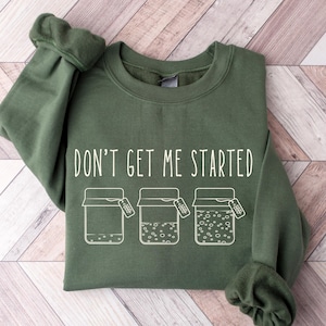 May include: An olive green sweatshirt with the phrase "DON'T GET ME STARTED" printed in white. Below the text are three jars with tags. The sweatshirt is made of a soft material and is laid out on a light brown and white wooden surface.