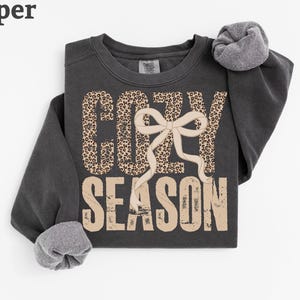 May include: A charcoal gray crewneck sweatshirt featuring the text COZY SEASON in a distressed beige font. The word COZY is filled with a brown and black leopard print pattern, and a large beige ribbon bow is centered over the letters.