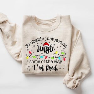 Peut inclure: Sweat-shirt beige avec l'inscription "Probably just gonna jingle some of the way. I'm tired." Le mot "Jingle" est surmont&eacute; d'un bonnet de P&egrave;re No&euml;l. Une guirlande de lumi&egrave;res de No&euml;l color&eacute;es est en dessous.