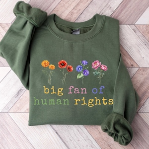 May include: Olive green sweatshirt with the text "big fan of human rights" in rainbow colors. The sweatshirt features a floral design with orange, red, purple, and pink flowers. The sleeves are cuffed.