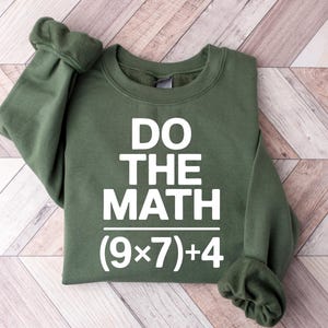 May include: An olive green sweatshirt with the text "DO THE MATH" in bold white letters. Below the text is the math equation (9x7)+4. The sweatshirt is folded, with the sleeves cuffed.