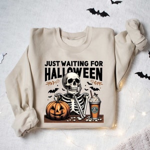 May include: A beige sweatshirt with the text "Just Waiting for Halloween" and a skeleton holding a pumpkin and a coffee drink.