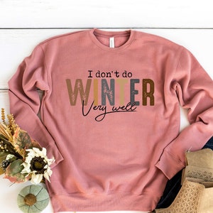 May include: A pink sweatshirt with the text "I don't do winter very well" in colorful letters. The sweatshirt is laid out on a white wooden surface with a bouquet of flowers and a pair of brown ankle boots.