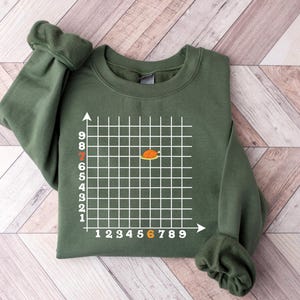 May include: An olive green sweatshirt featuring a white grid design with numbered axes. An orange and brown graphic is centered within the grid. The sweatshirt has ribbed cuffs and a crew neck.