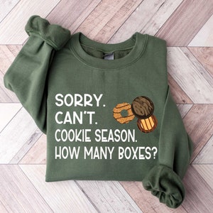 May include: Green sweatshirt with the text "SORRY. CAN'T. COOKIE SEASON. HOW MANY BOXES?" in white. There are three Girl Scout cookie images on the sweatshirt.