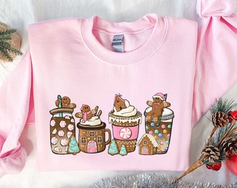 May include: Pink sweatshirt with a festive design featuring gingerbread men, candy canes, and hot chocolate. The design includes a gingerbread man wearing a Santa hat, a gingerbread man holding a candy cane, and a gingerbread man holding a cup of hot chocolate. The design also includes a gingerbread house and a Christmas tree.