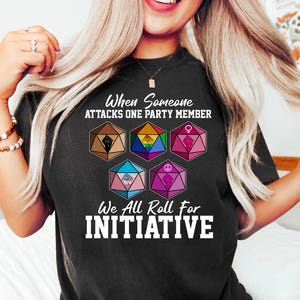 May include: Black t-shirt with the text "When Someone Attacks One Party Member, We All Roll For Initiative." The shirt features six multi-colored dice with various symbols, including a fist, rainbow, and gender symbols.