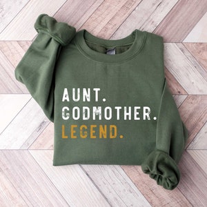 May include: An olive green sweatshirt with the words "AUNT. GODMOTHER. LEGEND." printed in white and gold. The sweatshirt is made of a soft material and has a crew neck and long sleeves. The text is centered on the front of the shirt.