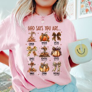 May include: A pink t-shirt with a graphic design featuring pumpkins, crosses, and bible verses. The text reads "God says you are..." and lists words like "chosen", "forgiven", "special", "lovely", "beautiful", "created", "unique", "strong", and "precious".