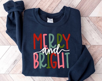 Op de afbeelding: Een marineblauw sweatshirt met de tekst "Merry and Bright" in kleurrijke letters. De letters zijn in rood, groen, roze en wit.