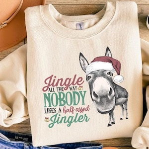 May include: Cream-colored sweatshirt with a graphic of a donkey wearing a Santa hat. Text reads "Jingle all the way, nobody likes a half assed Jingler." The sweatshirt is paired with blue jeans, a brown hat, and sandals.