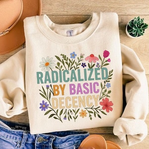 May include: Cream-colored sweatshirt with the text "RADICALIZED BY BASIC DECENCY" surrounded by colorful floral illustrations. The sweatshirt is paired with blue denim shorts, a brown hat, and sandals.