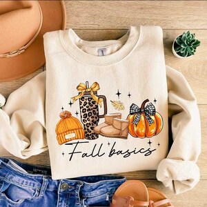 May include: Cream-colored sweatshirt with a fall-themed graphic. The graphic includes a leopard print tumbler, orange beanie, boots, and a pumpkin with a black and white checkered bow. The text "Fall basics" is written below the graphic.