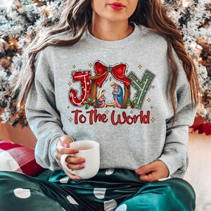 May include: A grey sweatshirt with a Christmas-themed design featuring the words "Joy to the World" and a depiction of the nativity scene. The design includes a red bow, green holly, and gold stars.