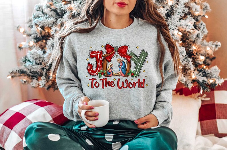 May include: A grey sweatshirt with a Christmas-themed design featuring the words "Joy to the World" and a depiction of the nativity scene. The design includes a red bow, green holly, and gold stars.