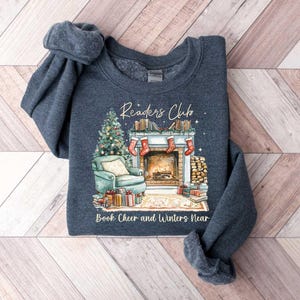K&ouml;nnte beinhalten: Ein dunkelblauer Pullover mit einer winterlichen Szene eines Kamins, B&uuml;chern und einem Weihnachtsbaum. Der Text "Readers Club" und "Book Cheer and Winters Near" ist auf dem Pullover aufgedruckt.