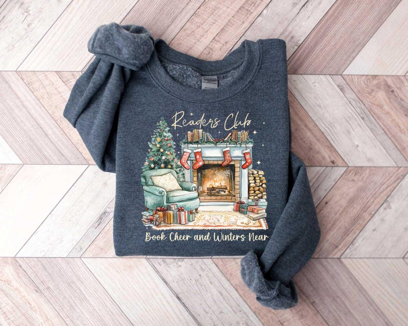 K&ouml;nnte beinhalten: Ein dunkelblauer Pullover mit einer winterlichen Szene eines Kamins, B&uuml;chern und einem Weihnachtsbaum. Der Text "Readers Club" und "Book Cheer and Winters Near" ist auf dem Pullover aufgedruckt.
