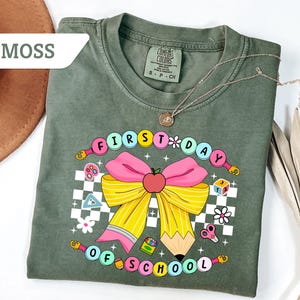 May include: A moss green t-shirt with a colorful graphic that says "FIRST DAY OF SCHOOL". The graphic includes a pink bow, an apple, a pencil, and other school-themed elements. The shirt is made of a soft material.