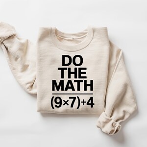 May include: A cream-colored sweatshirt with the text "DO THE MATH" in bold black letters. Below the text is the equation (9x7)+4. The sweatshirt is folded with the sleeves rolled up.