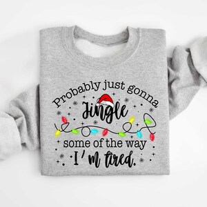 Peut inclure: Un sweat-shirt gris clair avec le texte "Probably just gonna Jingle some of the way. I'm tired." Le mot "Jingle" est surmont&eacute; d'un bonnet de P&egrave;re No&euml;l et entour&eacute; de guirlandes lumineuses.