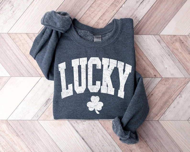 Może przedstawiać: Szara bluza z nadrukiem "LUCKY" w kolorze białym na przodzie. Słowo jest w dużym, pogrubionym kroju i wygięte nad białą koniczyną.