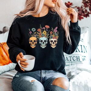 May include: A black sweatshirt with a floral skull design. The skulls are wearing floral crowns and have different colored eyes. The sweatshirt is being worn by a person with long blonde hair.