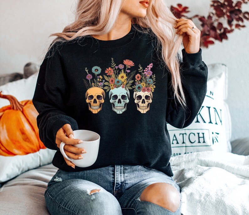 May include: A black sweatshirt with a floral skull design. The skulls are wearing floral crowns and have different colored eyes. The sweatshirt is being worn by a person with long blonde hair.