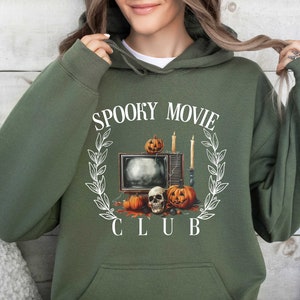 K&ouml;nnte beinhalten: Ein gr&uuml;ner Kapuzenpullover mit einem wei&szlig;en Grafikdesign eines Fernsehers mit K&uuml;rbissen und Kerzen. Der Text "Spooky Movie Club" ist um die Grafik gedruckt.