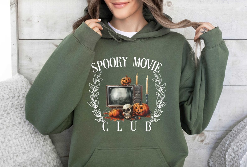 K&ouml;nnte beinhalten: Ein gr&uuml;ner Kapuzenpullover mit einem wei&szlig;en Grafikdesign eines Fernsehers mit K&uuml;rbissen und Kerzen. Der Text "Spooky Movie Club" ist um die Grafik gedruckt.