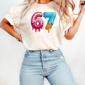 May include: Cream-colored t-shirt featuring the number "67" in a playful design. The "6" is a pink donut with sprinkles, and the "7" is a blue ice cream cone. The shirt is paired with light blue denim jeans.