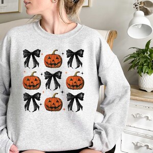 Op de afbeelding: Een grijs sweatshirt met een patroon van zwarte strikken en oranje pompoengezichten.