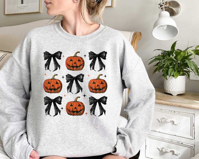 Op de afbeelding: Een grijs sweatshirt met een patroon van zwarte strikken en oranje pompoengezichten.