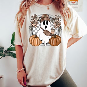 May include: A white t-shirt with a graphic of a ghost wearing a checkered hat, a brown crossbody bag, and holding a cup of coffee. The ghost is surrounded by orange pumpkins and brown flowers.