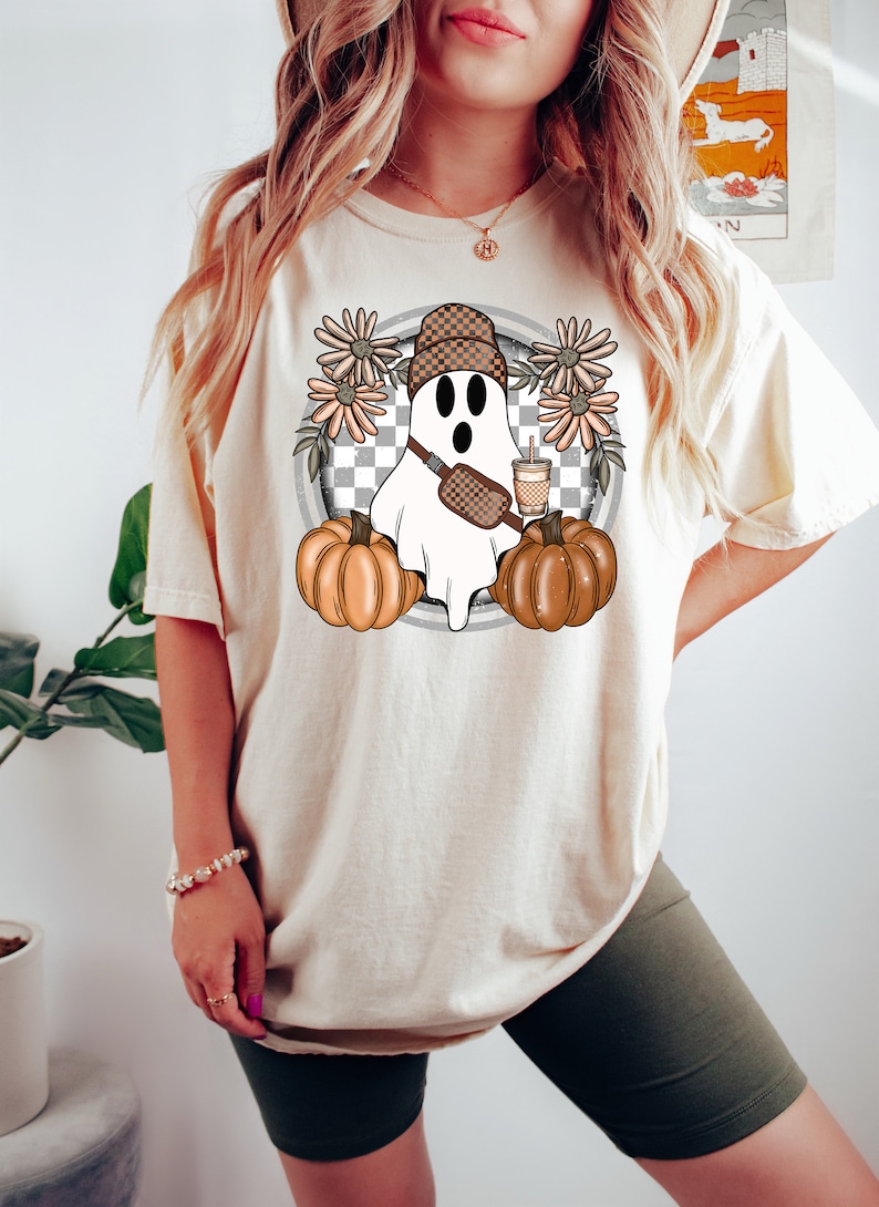 May include: A white t-shirt with a graphic of a ghost wearing a checkered hat, a brown crossbody bag, and holding a cup of coffee. The ghost is surrounded by orange pumpkins and brown flowers.