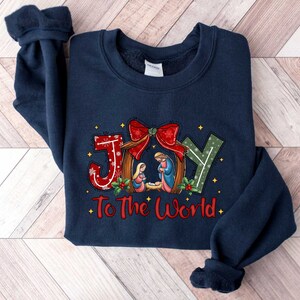 May include: Navy blue sweatshirt with a Christmas-themed design featuring the word "JOY" in red, green, and white letters with a red bow and a nativity scene. The text "To The World" is printed below the word "JOY".