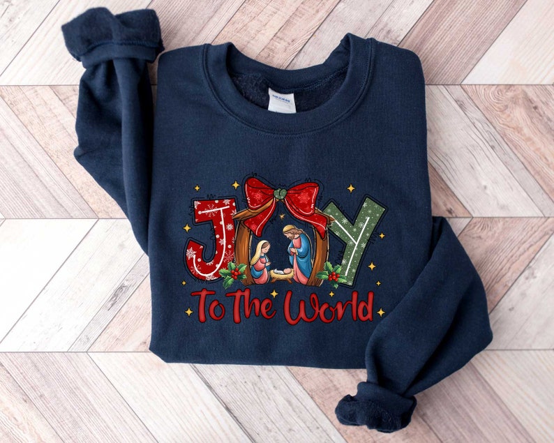May include: Navy blue sweatshirt with a Christmas-themed design featuring the word "JOY" in red, green, and white letters with a red bow and a nativity scene. The text "To The World" is printed below the word "JOY".