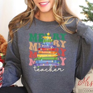 May include: A dark gray sweatshirt with a colorful Christmas tree design. The tree is made of stacked books and the words "MERRY MERRY MERRY MERRY" are above it. The word "teacher" is written below the tree in cursive.