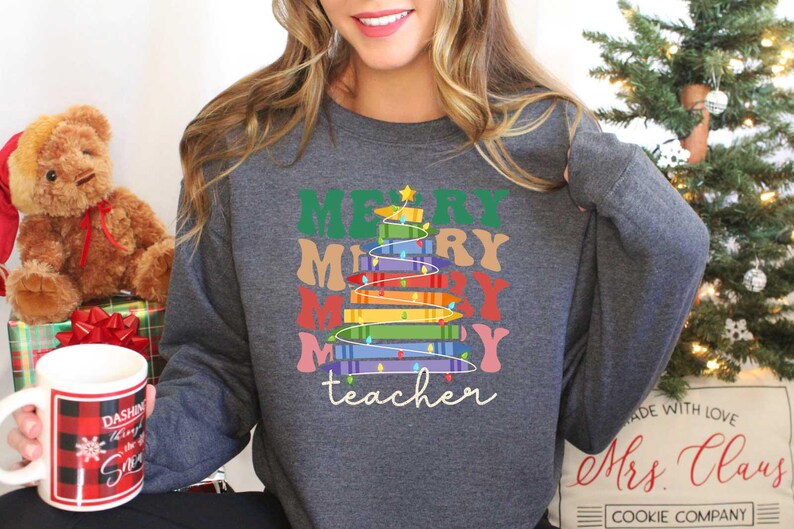 May include: A dark gray sweatshirt with a colorful Christmas tree design. The tree is made of stacked books and the words "MERRY MERRY MERRY MERRY" are above it. The word "teacher" is written below the tree in cursive.
