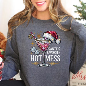 May include: A gray sweatshirt with a sparkly pink martini glass with a Santa hat on top. The text "Santa's Favorite Hot Mess" is printed on the sweatshirt.