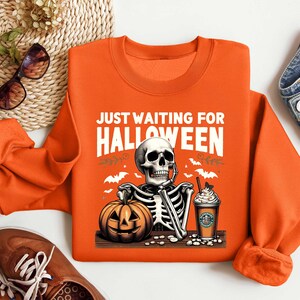 May include: Orange sweatshirt with a skeleton holding a pumpkin and a cup of coffee. The text on the sweatshirt says "Just Waiting for Halloween".