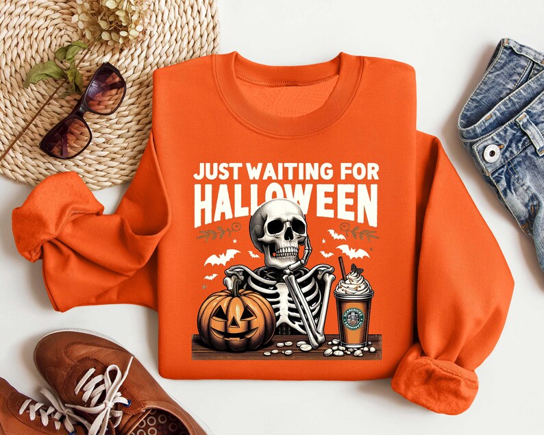 May include: Orange sweatshirt with a skeleton holding a pumpkin and a cup of coffee. The text on the sweatshirt says "Just Waiting for Halloween".