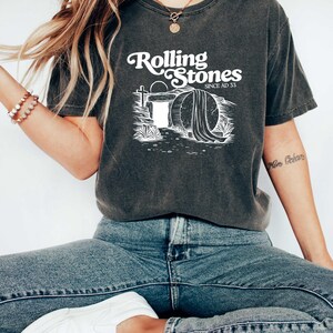 May include: A dark gray t-shirt with a white graphic of a stone tomb with the text "Rolling Stones Since AD 33".