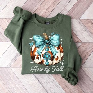 Puede incluir: Sudadera verde con una calabaza de estampado de vaca marr&oacute;n y blanco con un lazo turquesa y pedrer&iacute;a. El texto "Howdy Fall" est&aacute; impreso debajo de la calabaza.