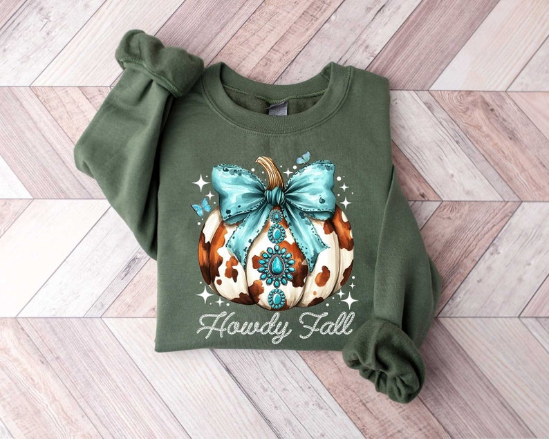 Puede incluir: Sudadera verde con una calabaza de estampado de vaca marr&oacute;n y blanco con un lazo turquesa y pedrer&iacute;a. El texto "Howdy Fall" est&aacute; impreso debajo de la calabaza.