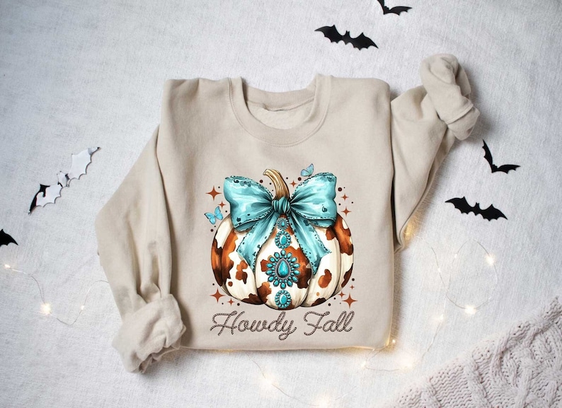 Puede incluir: Una sudadera beige con una calabaza de estampado de vaca marr&oacute;n y blanco con un lazo turquesa y pedrer&iacute;a. El texto "Howdy Fall" est&aacute; impreso debajo de la calabaza.