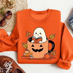 May include: Orange sweatshirt featuring a ghost reading a book inside a pumpkin mug. The ghost is white with a smiling face and the pumpkin mug has a jack-o'-lantern face. There are autumn leaves around the mug.