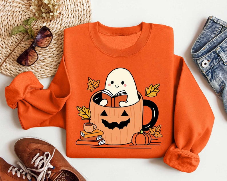 May include: Orange sweatshirt featuring a ghost reading a book inside a pumpkin mug. The ghost is white with a smiling face and the pumpkin mug has a jack-o'-lantern face. There are autumn leaves around the mug.