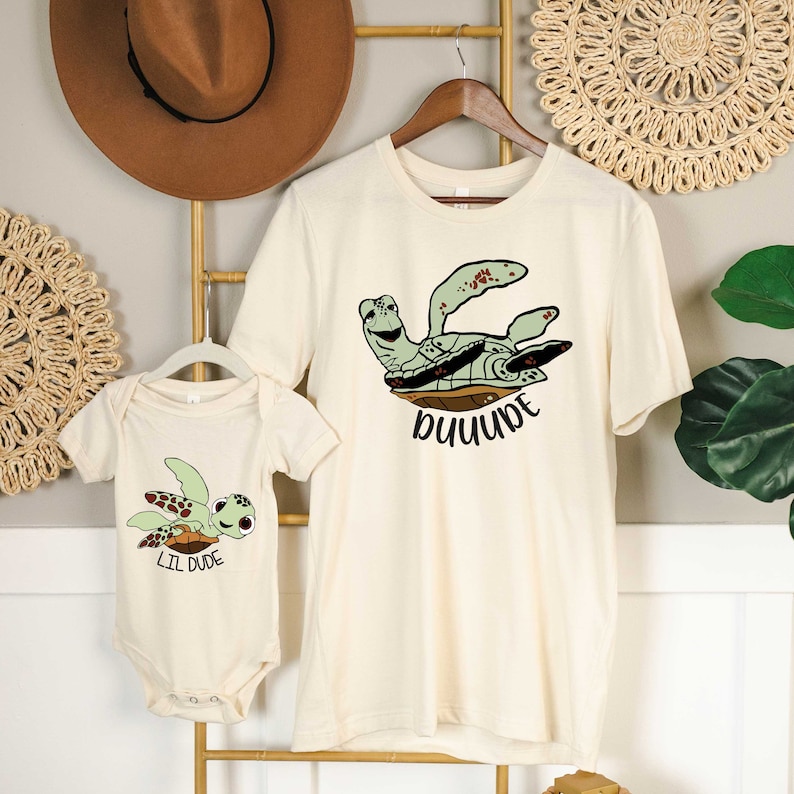 May include: A set of two cream coloured shirts featuring a cartoon sea turtle design. The larger shirt says "DUDE" and the smaller shirt says "LIL DUDE".