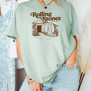 May include: A light green t-shirt with a brown graphic of a tomb with a stone rolled away and the text "Rolling Stones Since AD 33".