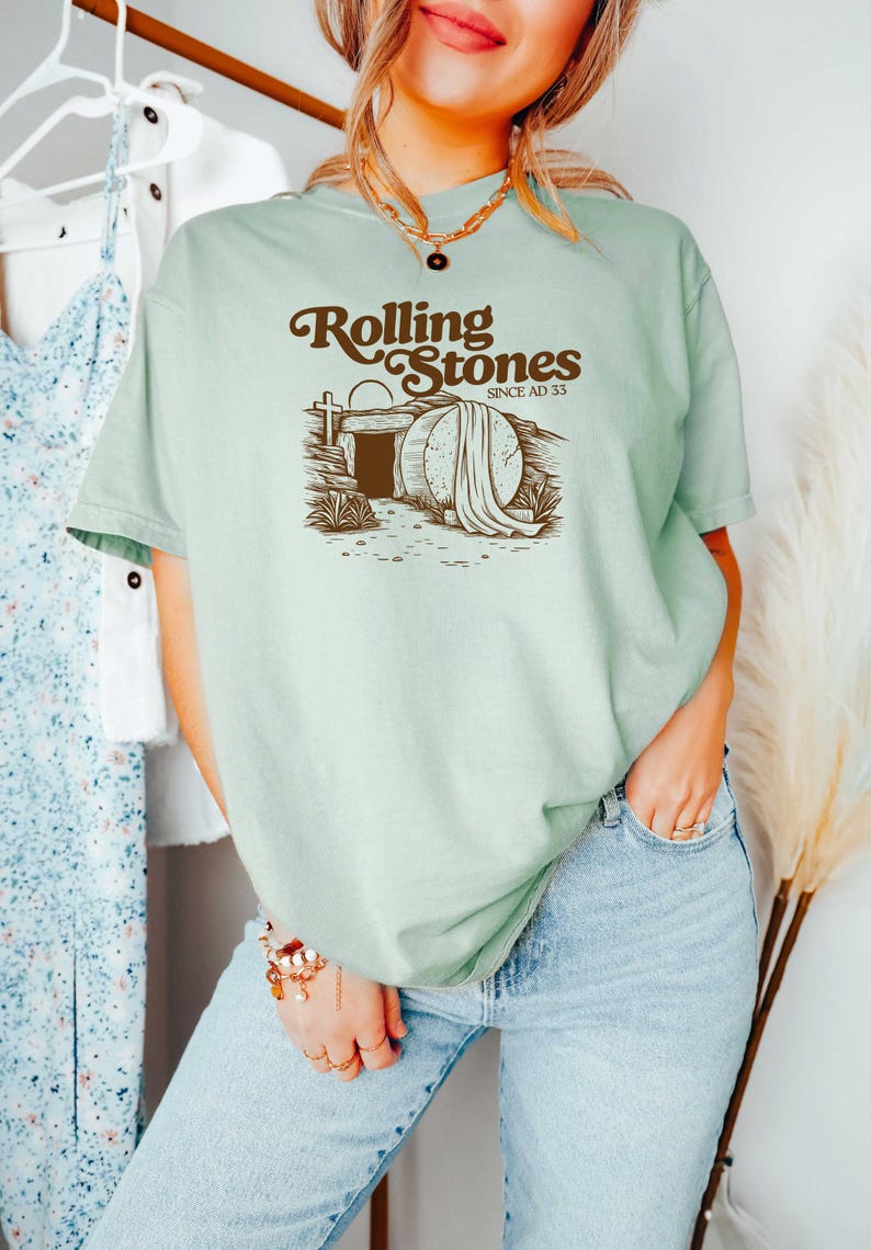 May include: A light green t-shirt with a brown graphic of a tomb with a stone rolled away and the text "Rolling Stones Since AD 33".